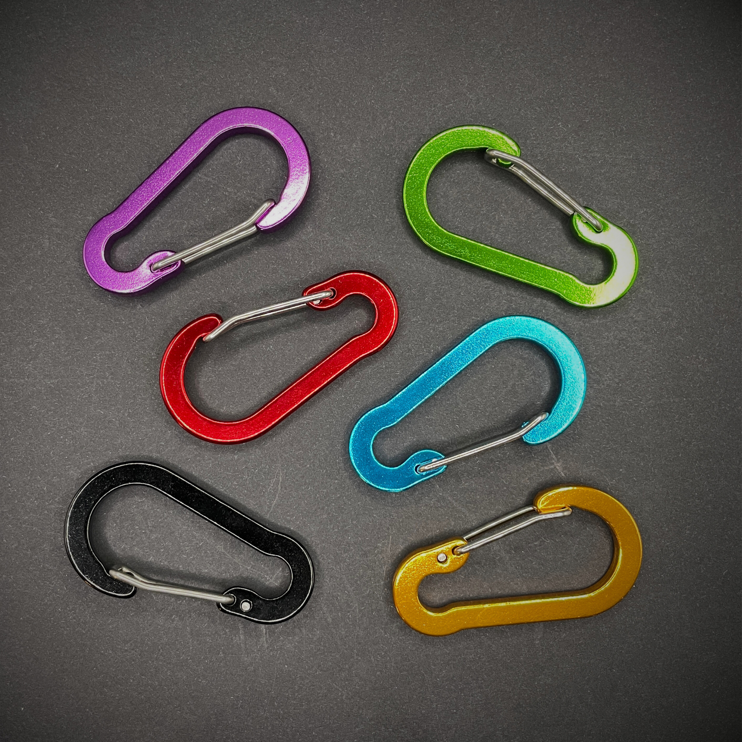 Six colourful aluminium carabiners and arranged on a dark surface.