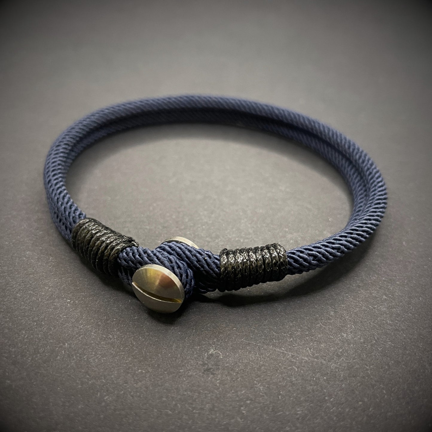 Close-up of a navy rope bracelet with a screw clasp and black whipping detail, displayed on a dark surface.