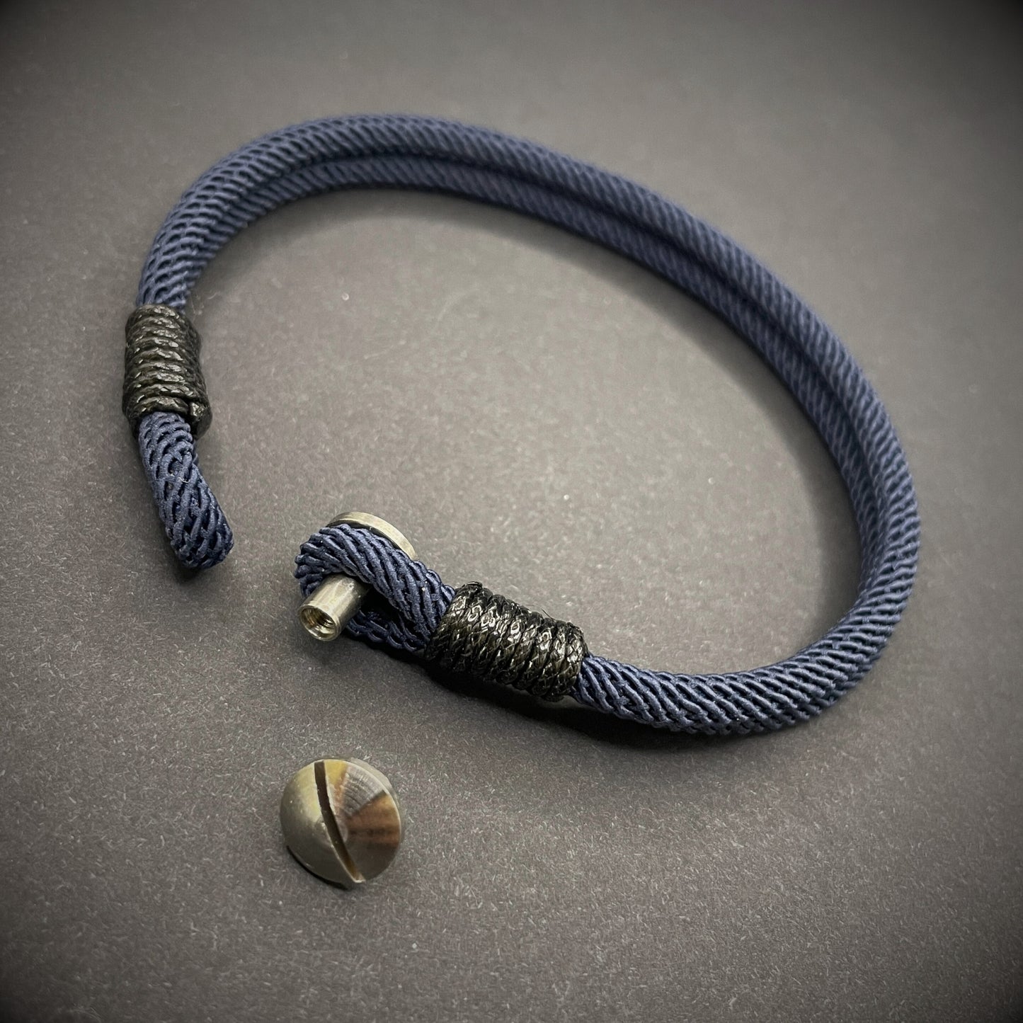 Close-up of a navy rope bracelet with a screw clasp and black whipping detail, displayed on a dark surface.