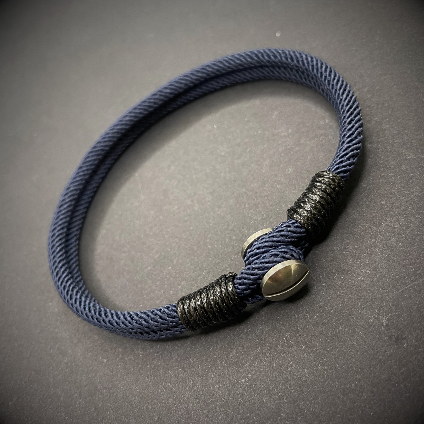 Close-up of a navy rope bracelet with a screw clasp and black whipping detail, displayed on a dark surface.