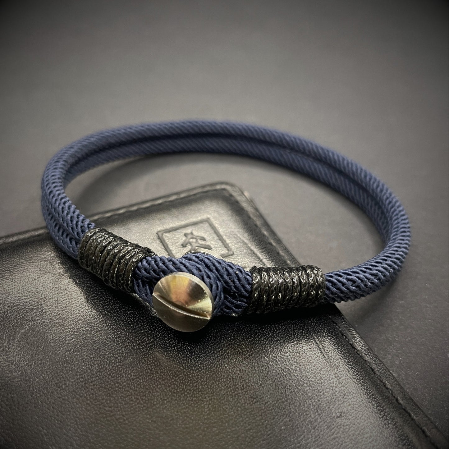 Close-up of a navy rope bracelet with a screw clasp and black whipping detail, displayed on a dark surface.