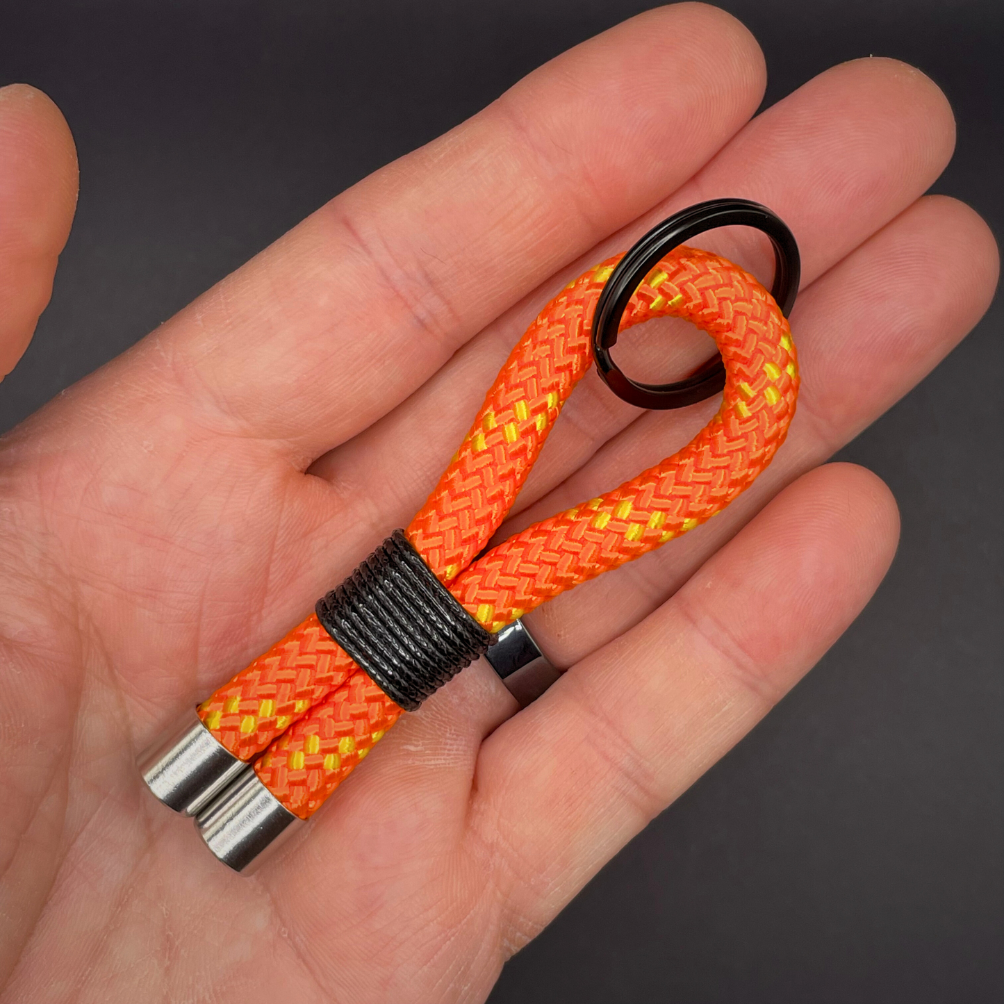 Flat lay of black braided rope keychain with silver end caps and orange whipping on a dark background.