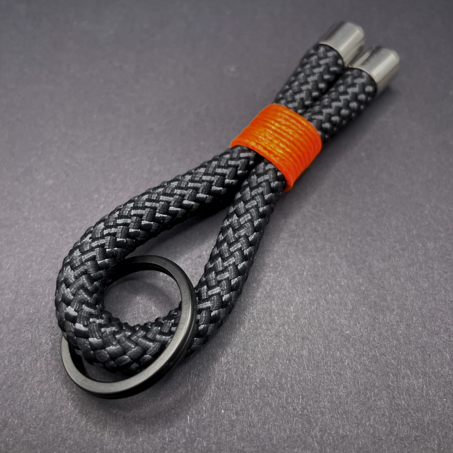 Flat lay of black braided rope keychain with silver end caps and orange whipping on a dark background.