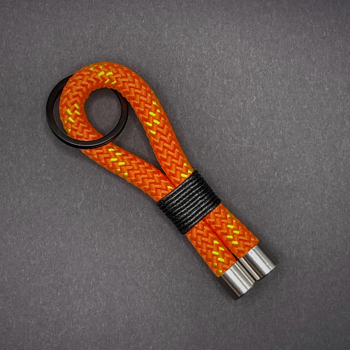 Flat lay of black braided rope keychain with silver end caps and orange whipping on a dark background.