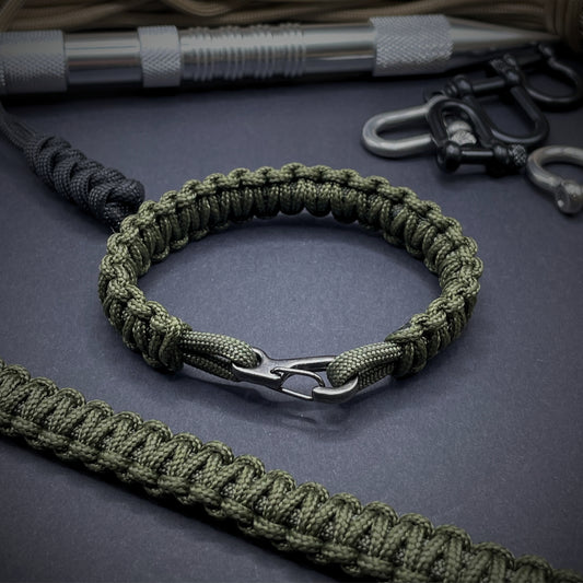 Flat lay of Army Green cobra knot paracord bracelet on a dark grey surface surrounded by EDC items