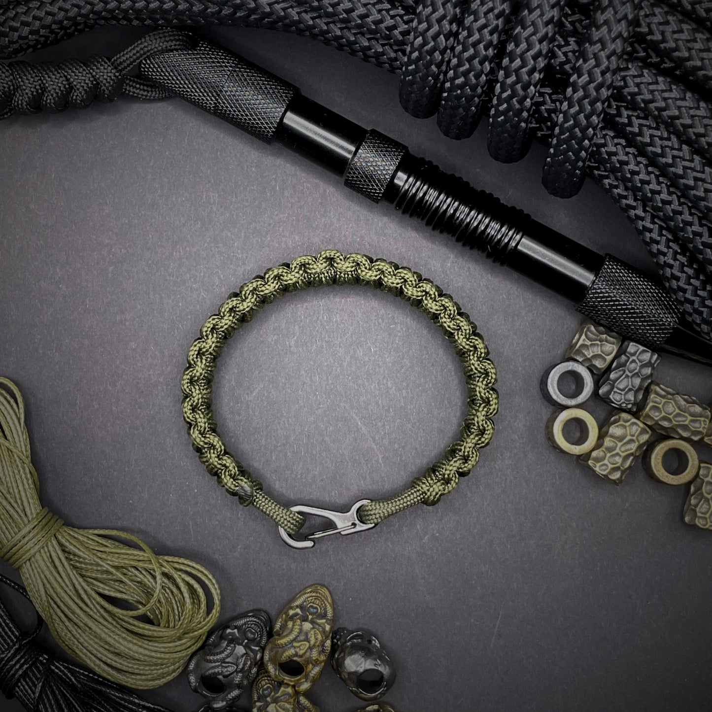 Flat lay of Army Green cobra knot paracord bracelet on a dark grey surface surrounded by EDC items