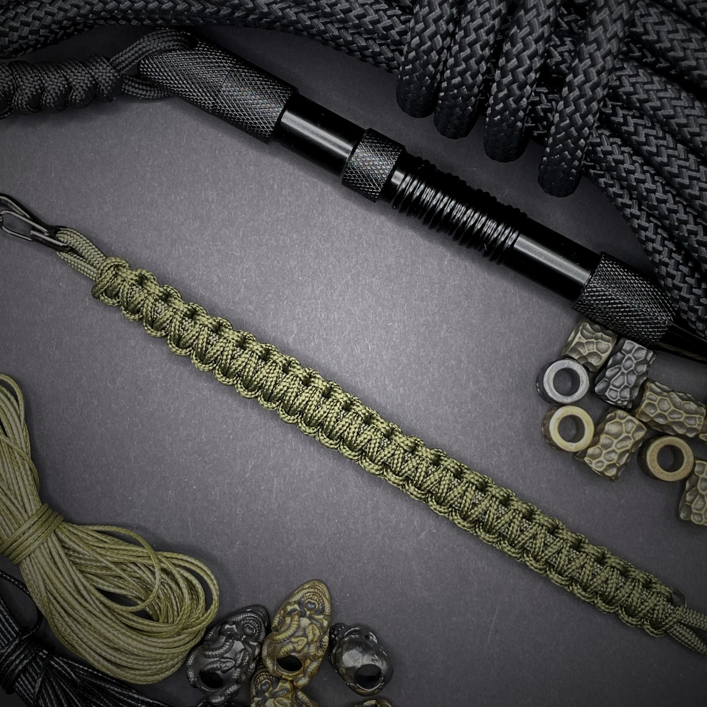 Flat lay of Army Green cobra knot paracord bracelet on a dark grey surface surrounded by EDC items