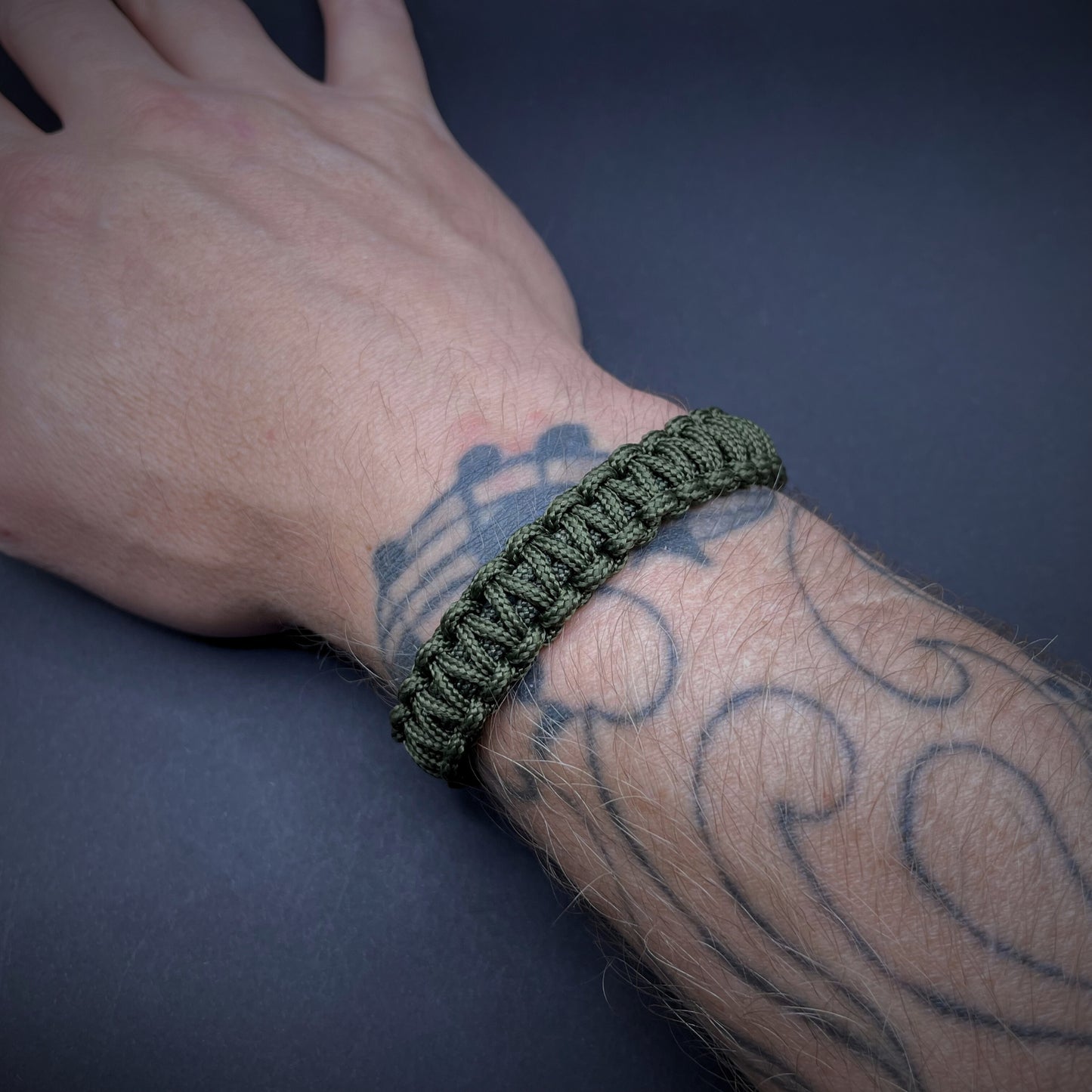 Army Green cobra knot paracord bracelet shown on a tattooed wrist over a dark grey surface