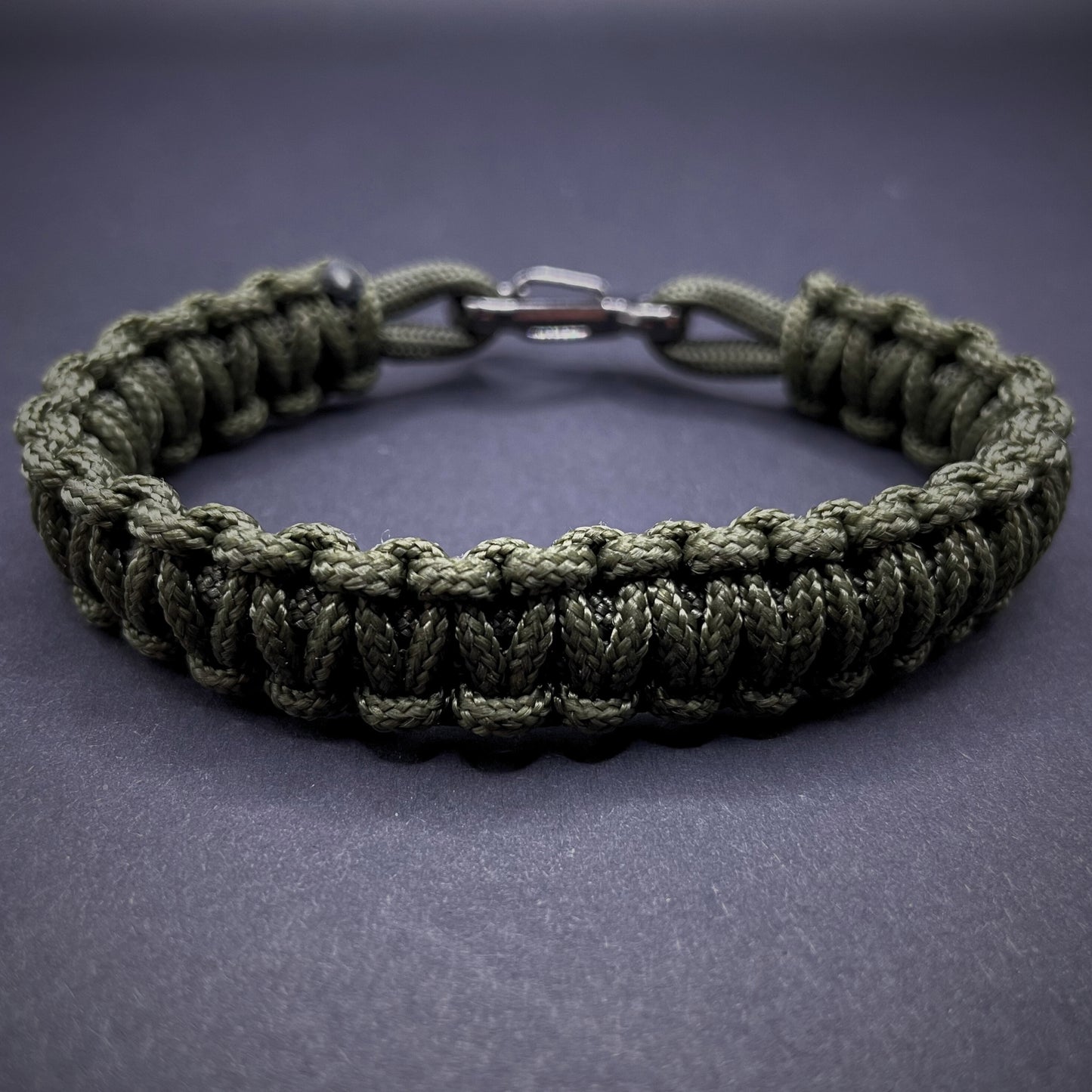 Flat lay of Army Green cobra knot paracord bracelet on a dark grey surface surrounded by EDC items