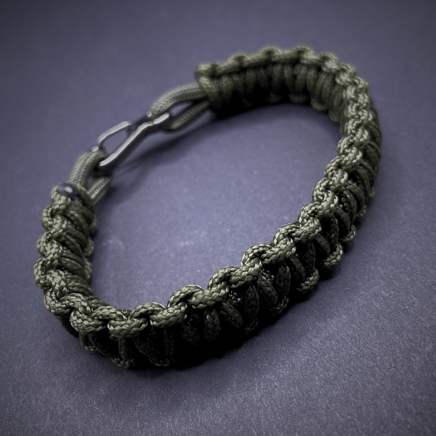 Flat lay of Army Green cobra knot paracord bracelet on a dark grey surface surrounded by EDC items