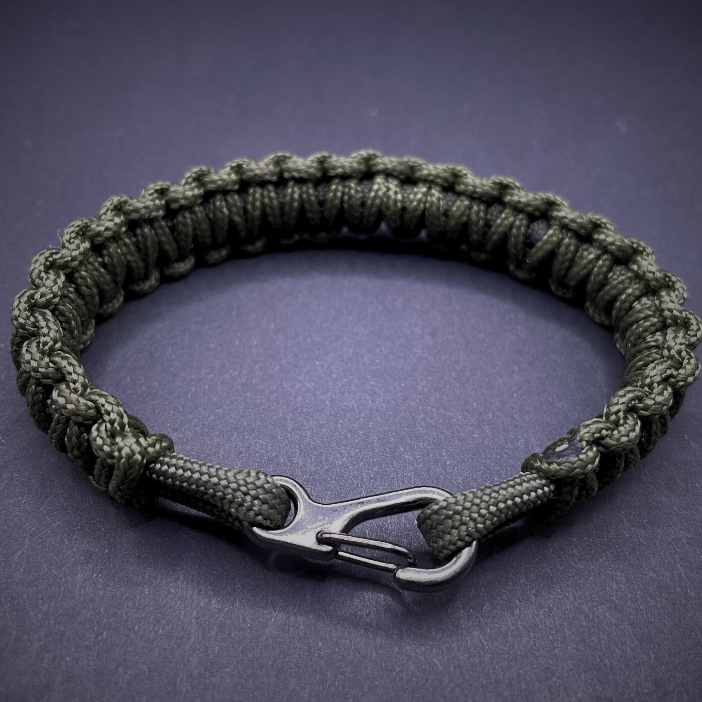 Flat lay of Army Green cobra knot paracord bracelet on a dark grey surface surrounded by EDC items