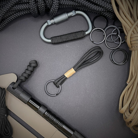 Flat lay of black 550 paracord keychain with tan cotton whipping on a dark background.