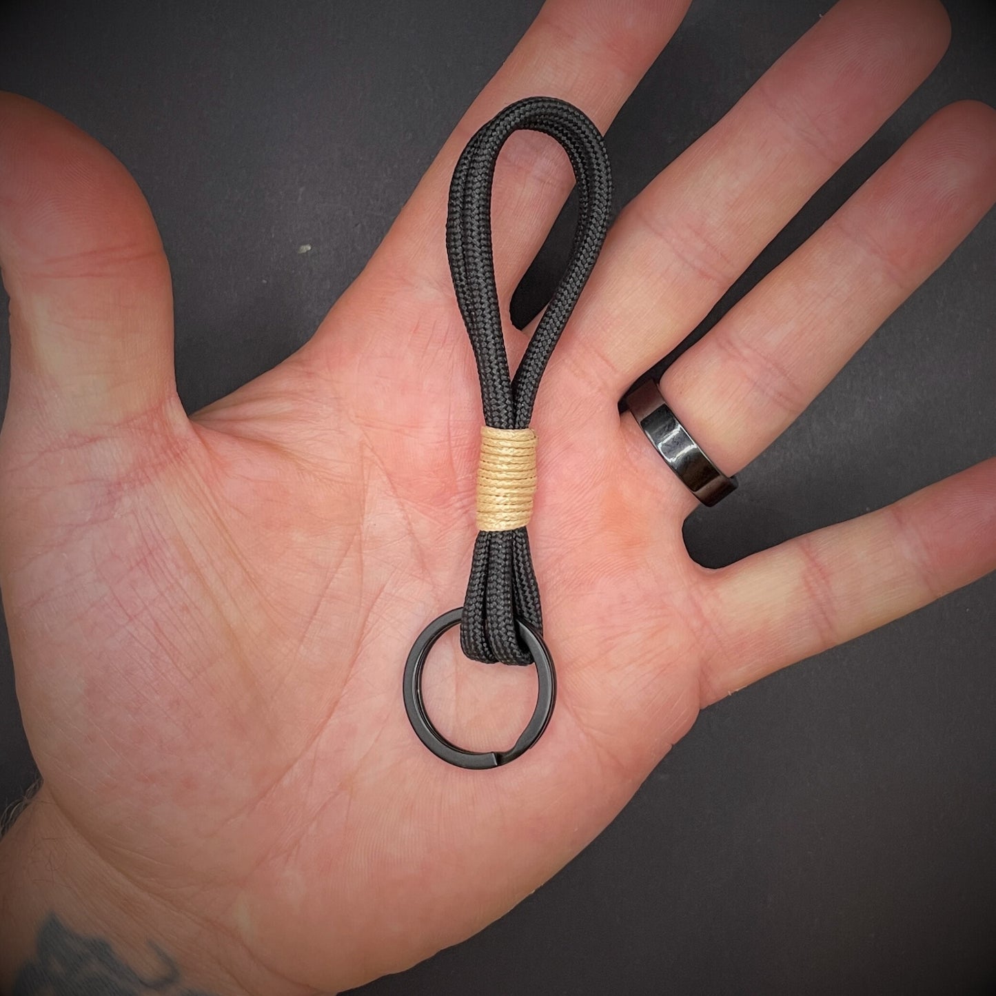 Flat lay of black 550 paracord keychain with tan cotton whipping on a dark background.