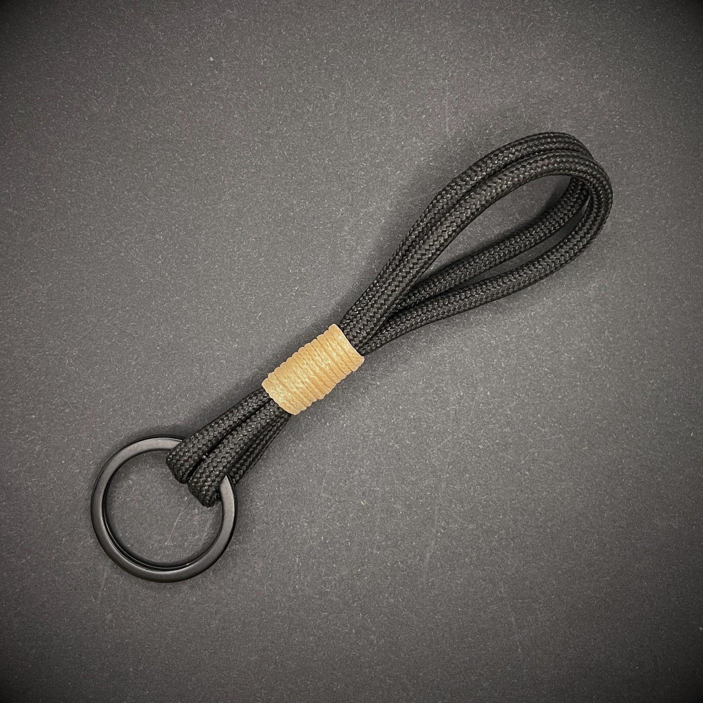 Flat lay of black 550 paracord keychain with tan cotton whipping on a dark background.