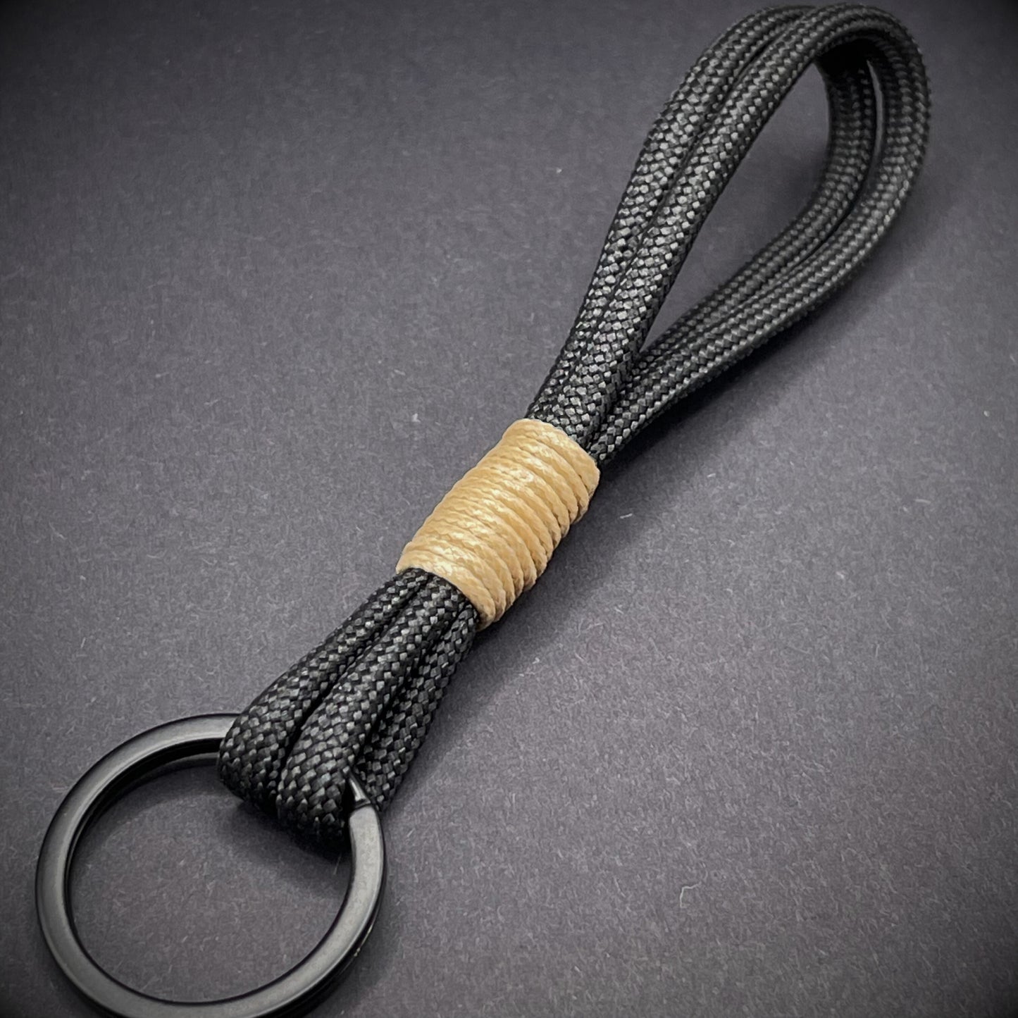 Flat lay of black 550 paracord keychain with tan cotton whipping on a dark background.