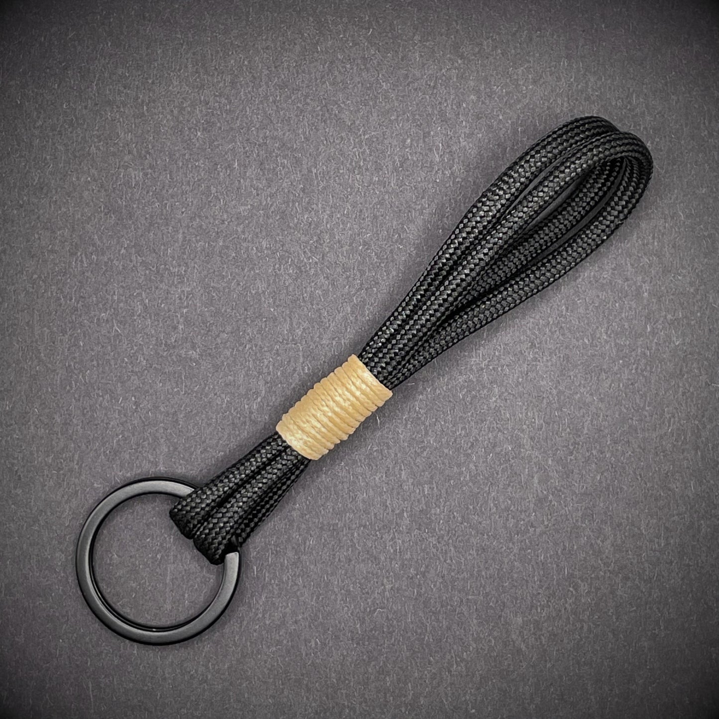 Flat lay of black 550 paracord keychain with tan cotton whipping on a dark background.