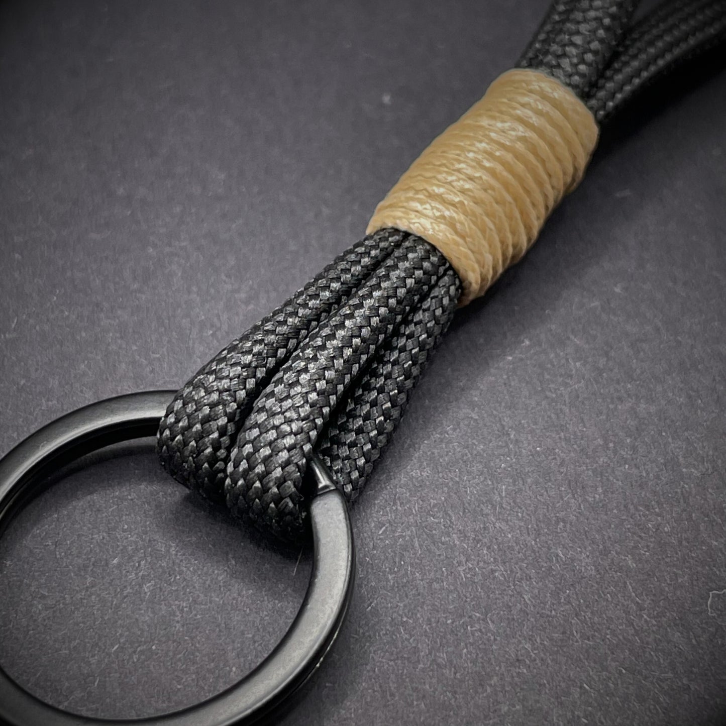 Flat lay of black 550 paracord keychain with tan cotton whipping on a dark background.