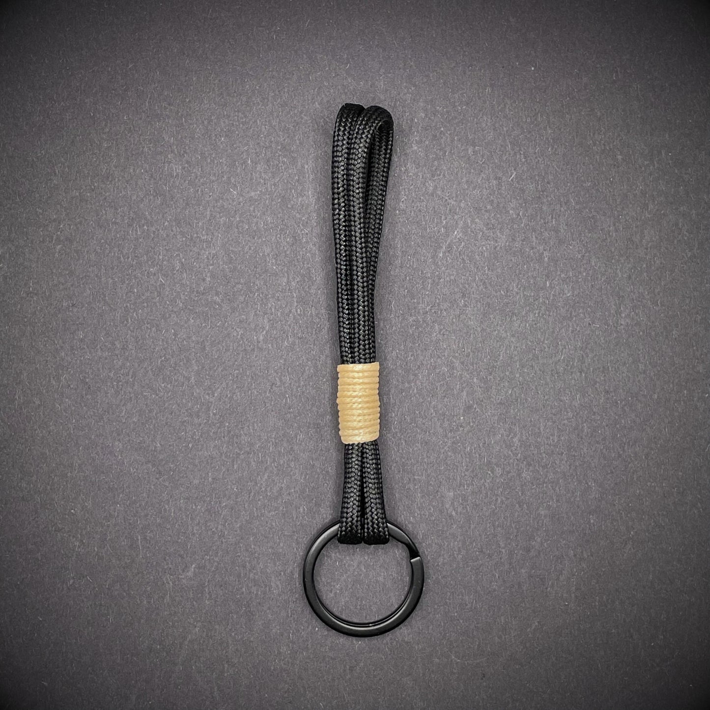 Flat lay of black 550 paracord keychain with tan cotton whipping on a dark background.