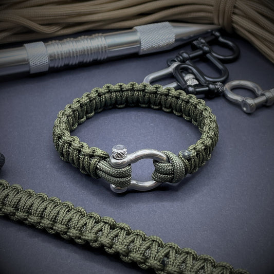 Flat lay of Army Green cobra knot paracord bracelet on a dark grey surface surrounded by EDC items