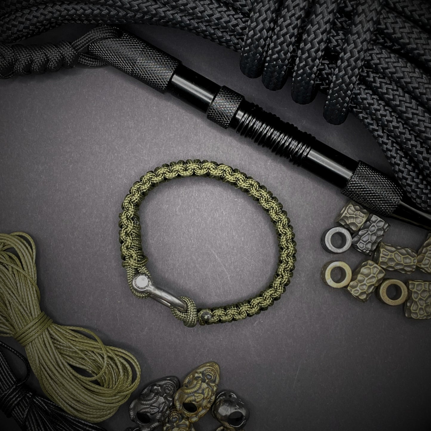 Flat lay of Army Green cobra knot paracord bracelet on a dark grey surface surrounded by EDC items