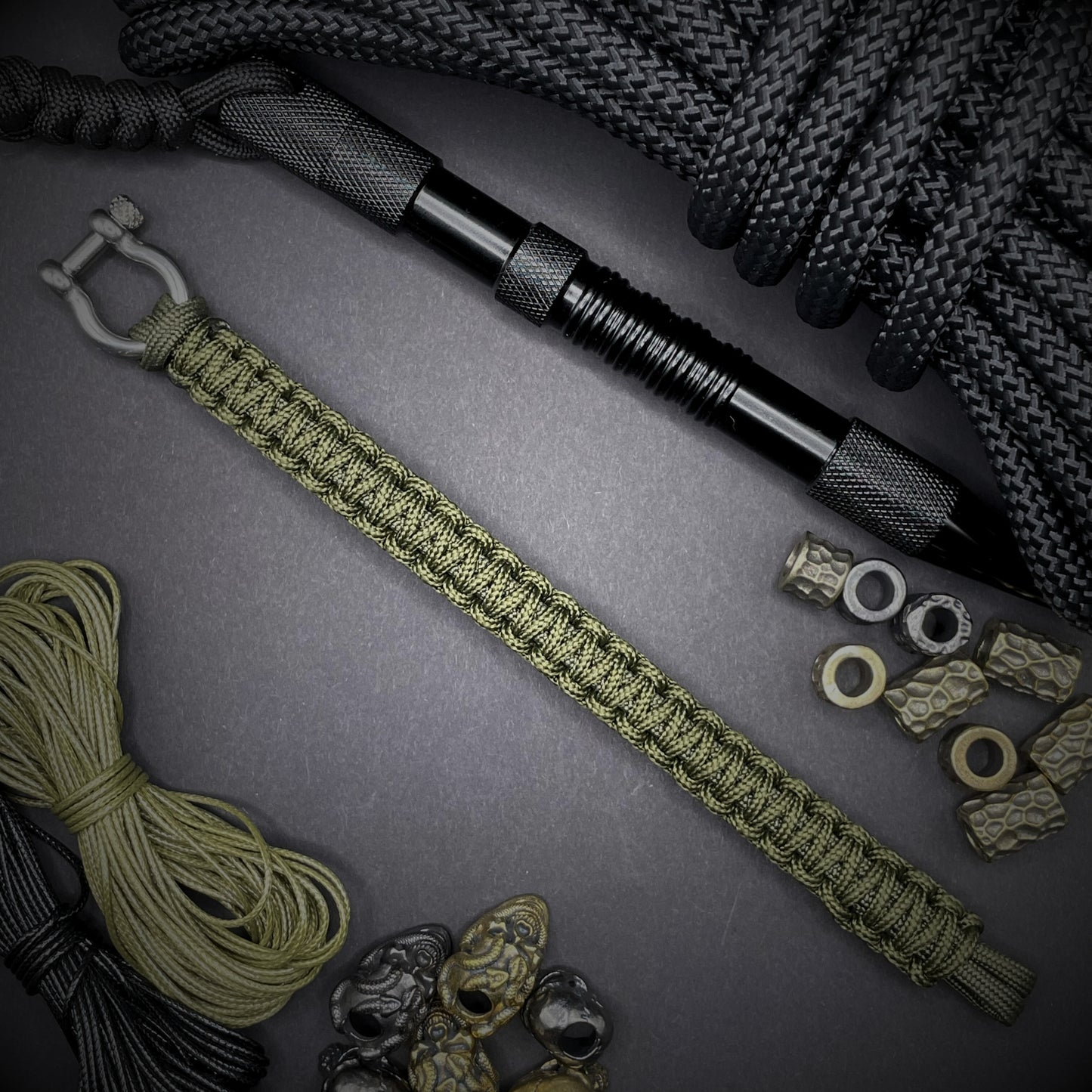 Flat lay of Army Green cobra knot paracord bracelet on a dark grey surface surrounded by EDC items