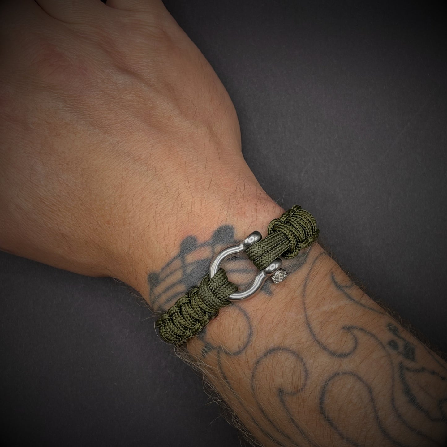 Flat lay of Army Green cobra knot paracord bracelet on a dark grey surface surrounded by EDC items