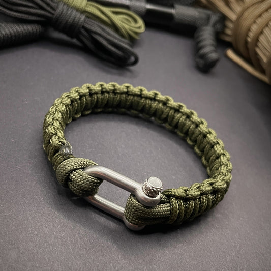 An army green paracord rope bracelet arranged on a dark grey surface with EDC gear in the background.