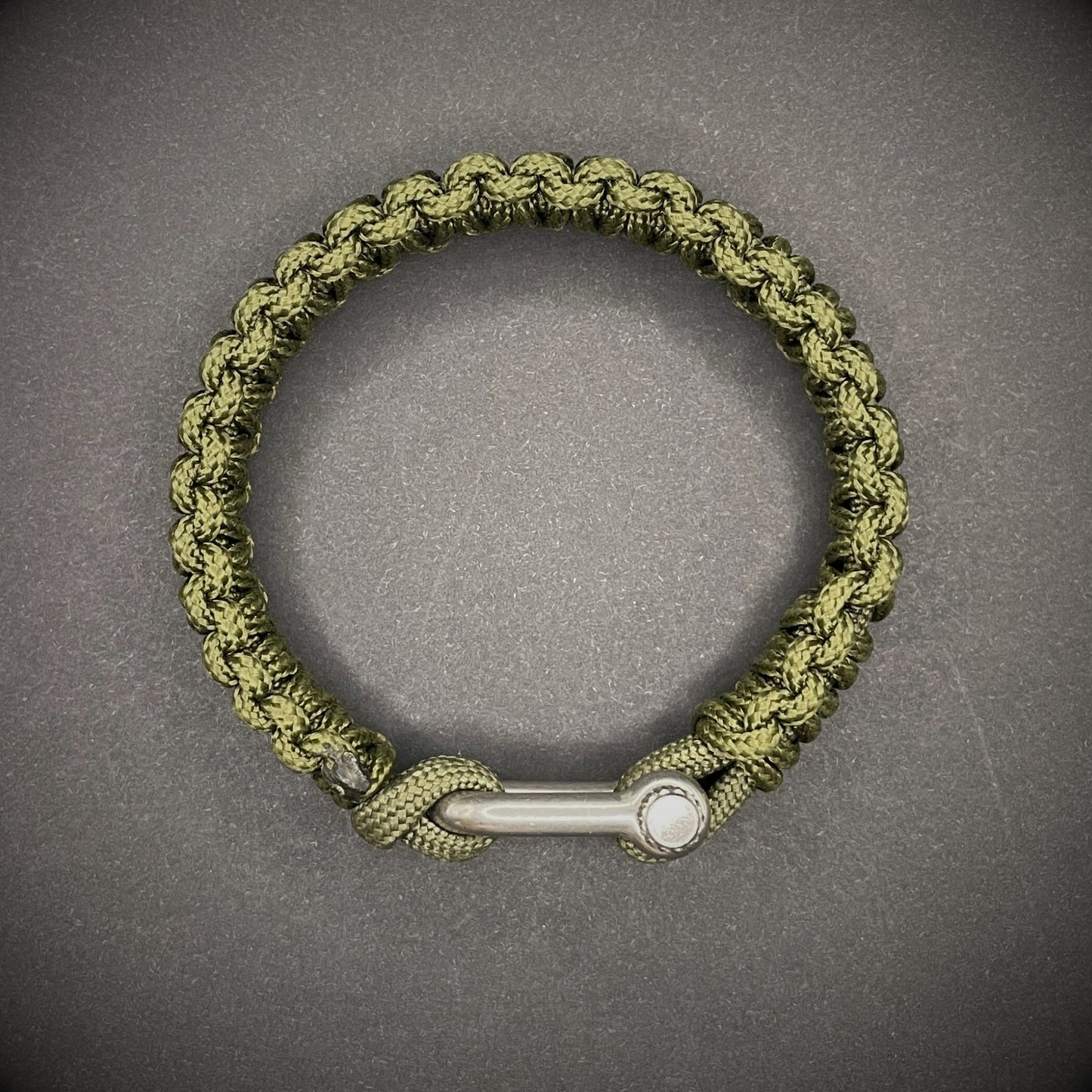 An army green paracord rope bracelet arranged on a dark grey surface.