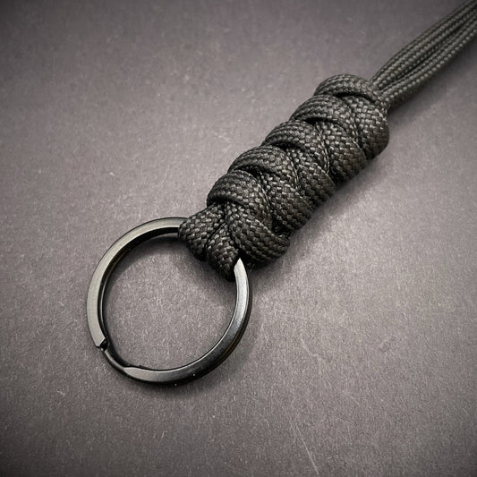 Black 550 Paracord snake knot lanyard with black split ring, displayed on a dark textured background.