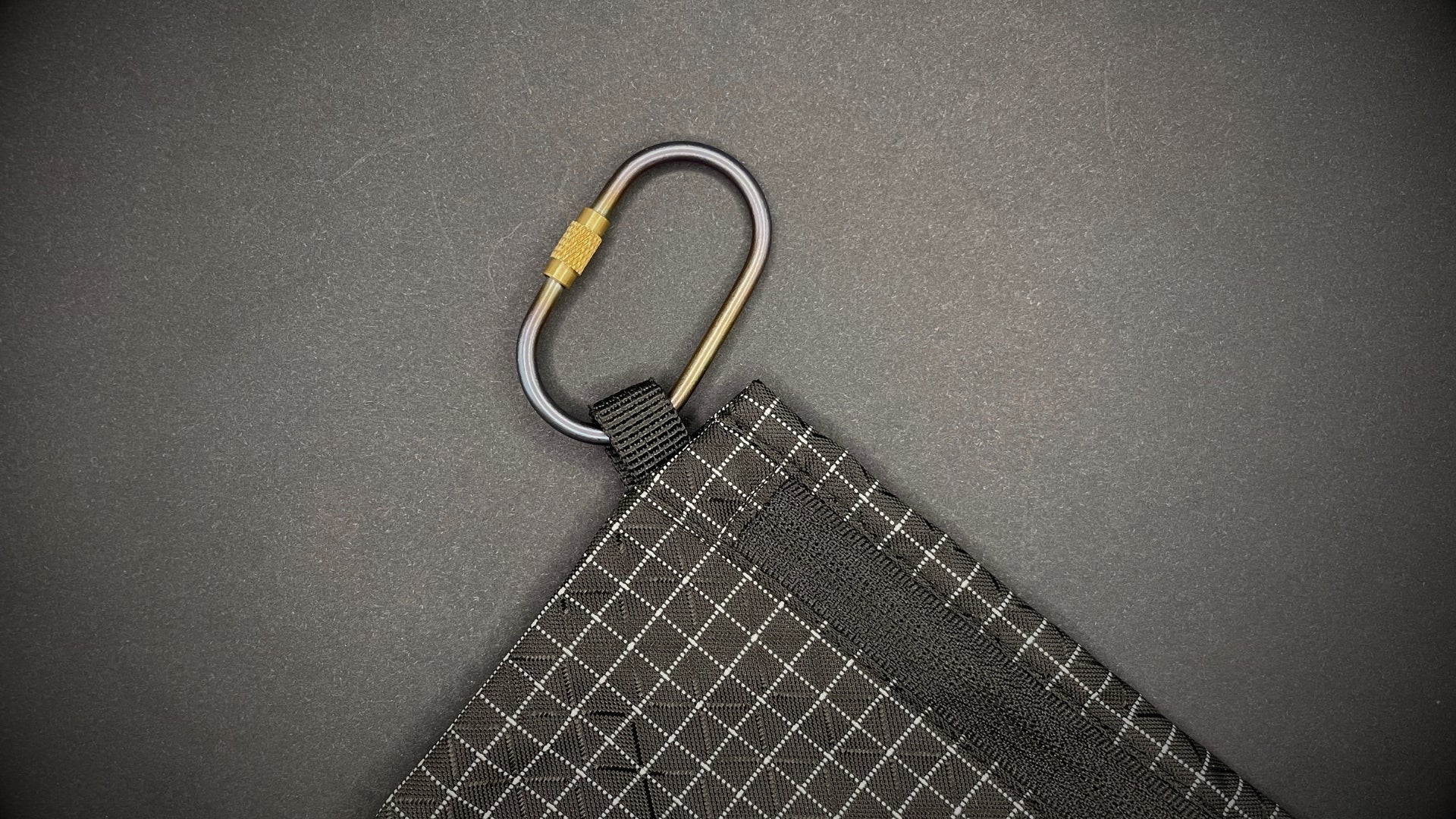 Mini Locking Titanium Carabiner attached to a black grid-patterned pouch, arranged on a dark surface.