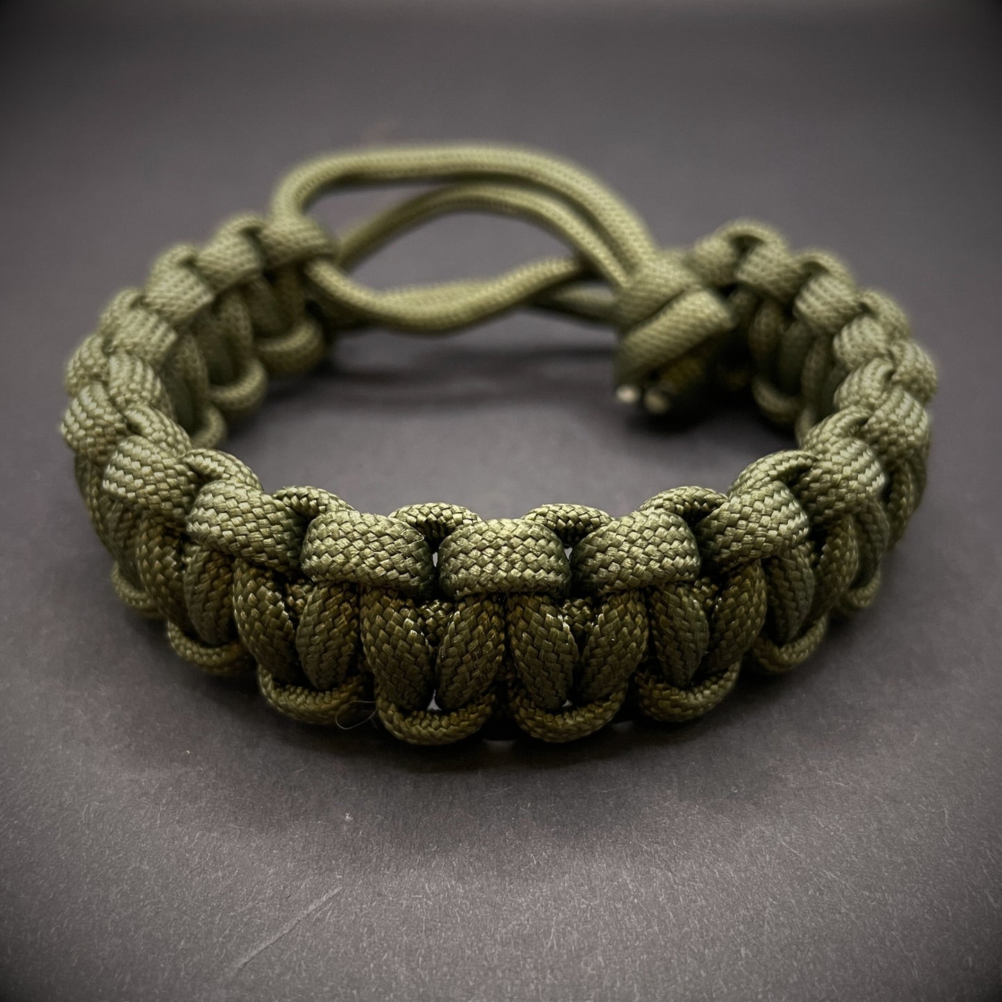 An Army green 550 paracord cobra knot bracelet displayed on a dark grey surface.