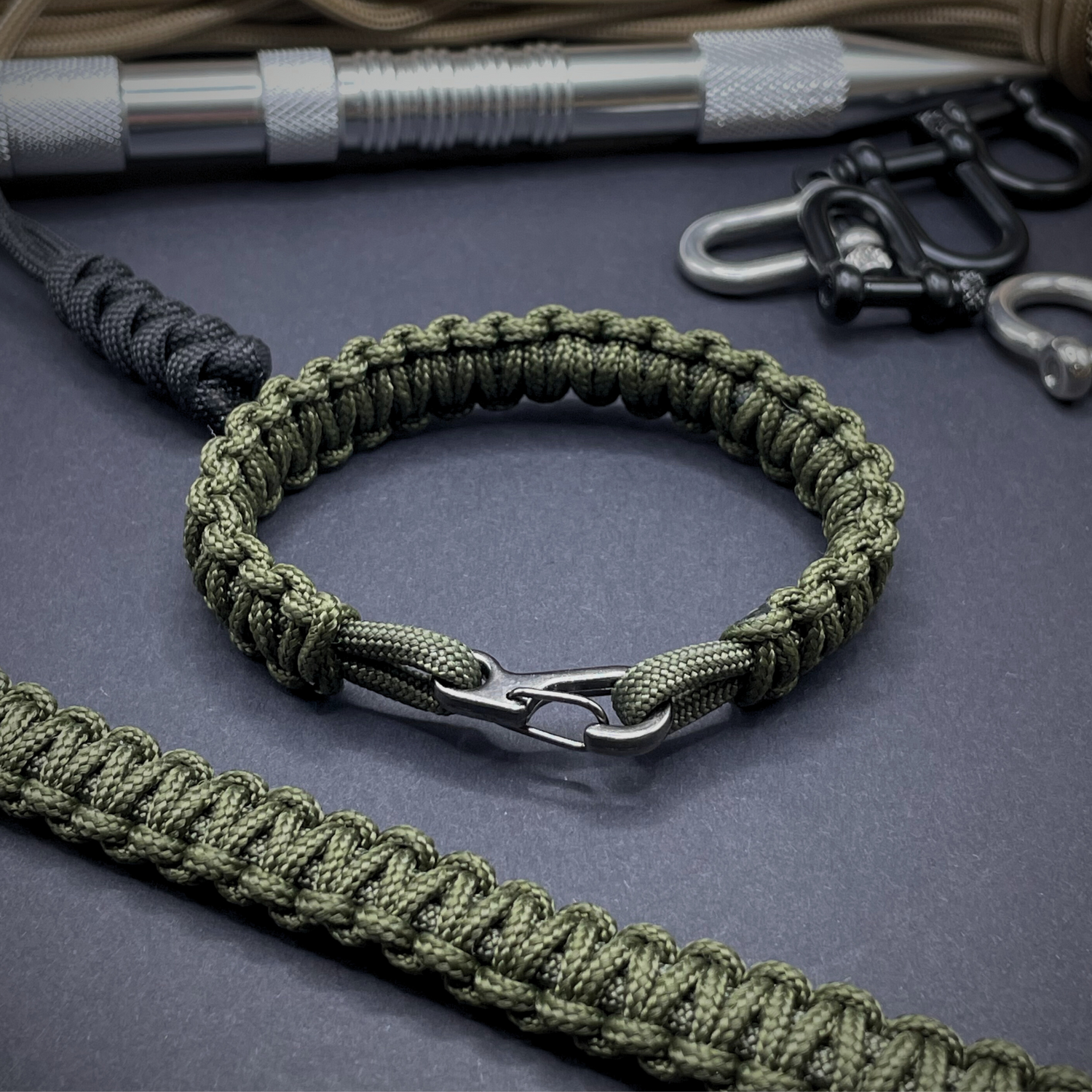 Army green paracord bracelet with a metal clasp, surrounded by tactical gear, including a marlinspike, clips, and braided cord.