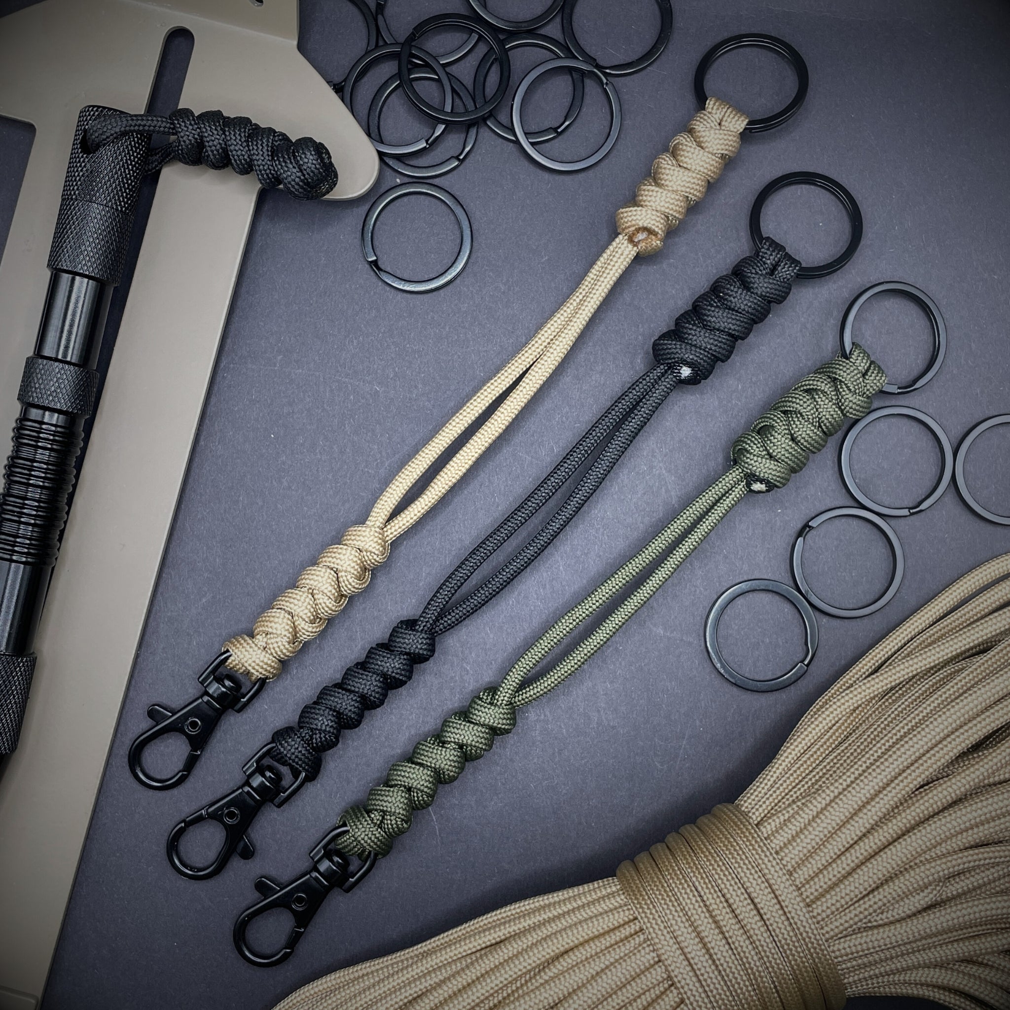 Three 550 Paracord snake knot lanyards with black split ring and lobster clasp, displayed on a dark textured background with EDC gear in the background.