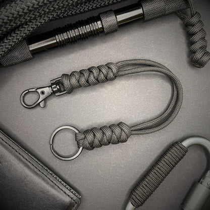 Black 550 Paracord snake knot lanyard with black split ring and lobster clasp, displayed on a dark textured background with EDC gear in the background.