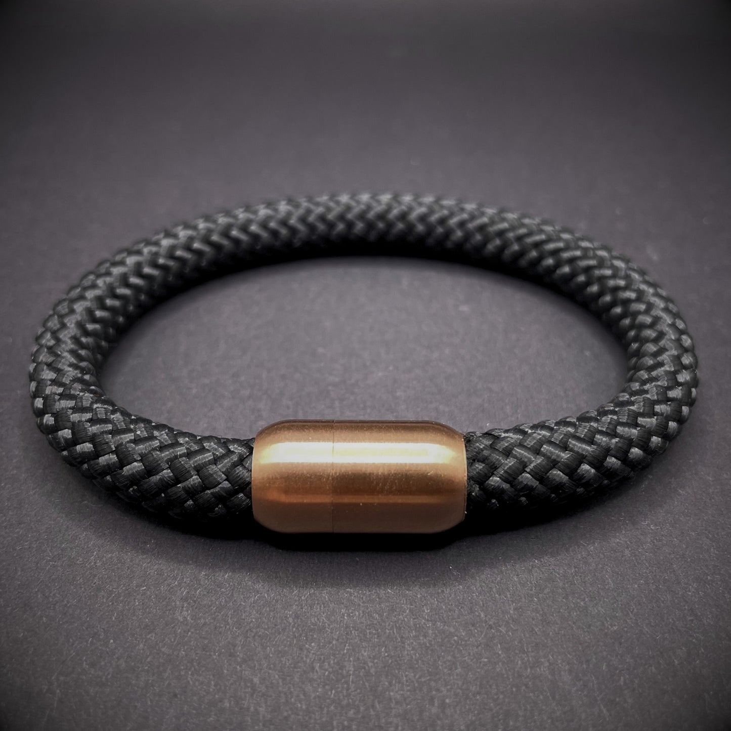 Black braided rope bracelet with a sleek cylindrical rose gold metal clasp, displayed on a dark textured background.