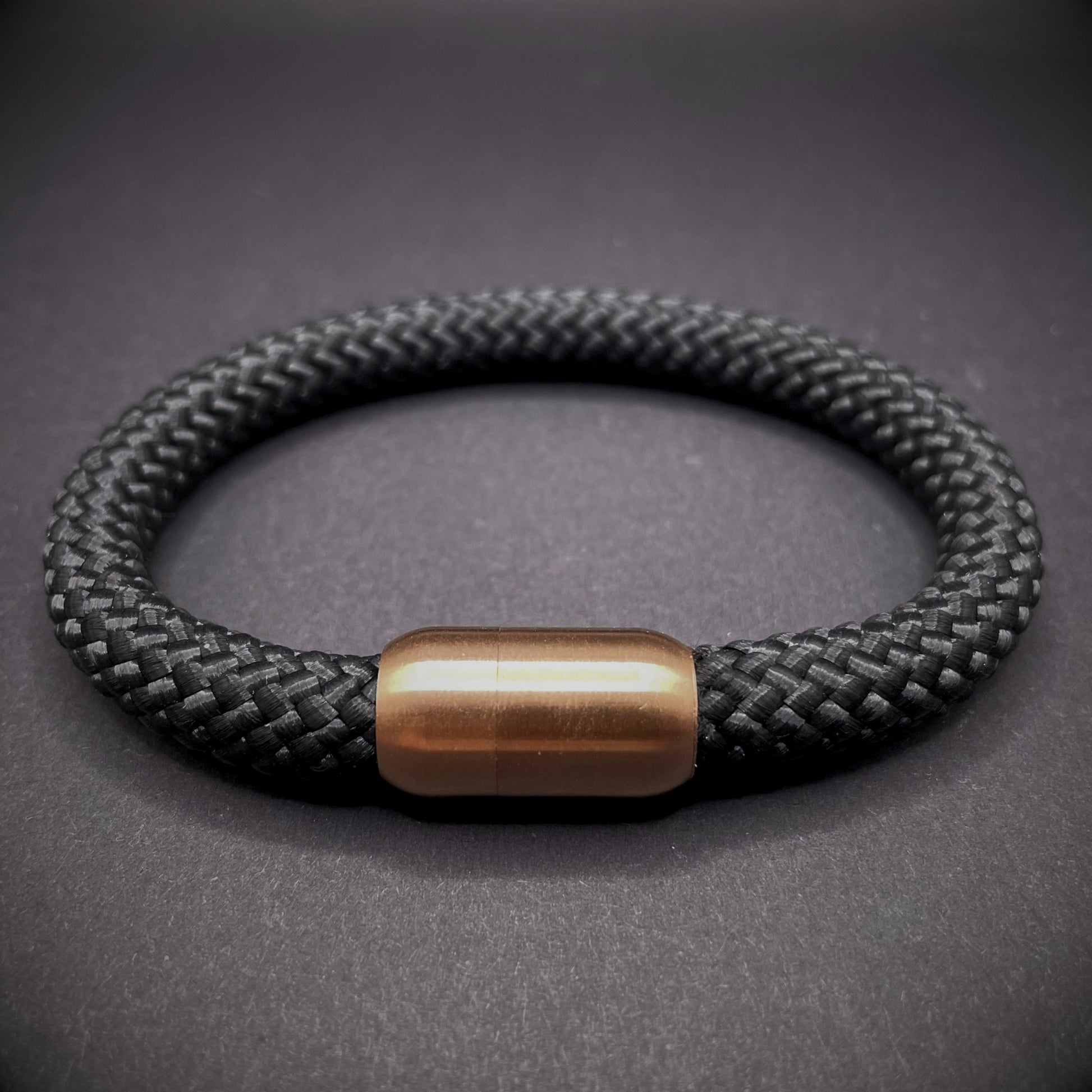 Black braided rope bracelet with a sleek cylindrical rose gold metal clasp, displayed on a dark textured background.