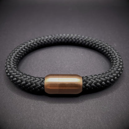Black braided rope bracelet with a sleek cylindrical rose gold metal clasp, displayed on a dark textured background.