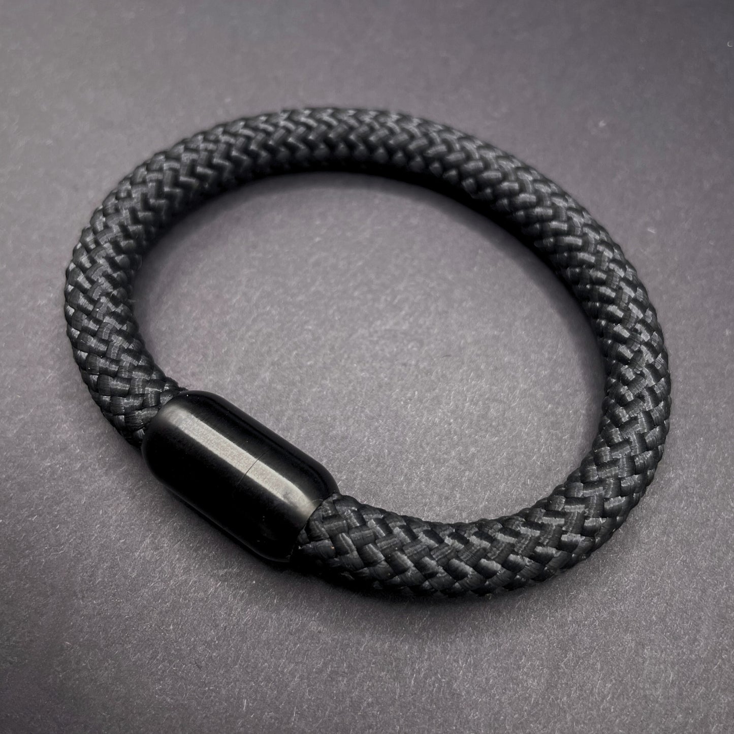 Black braided rope bracelet with a sleek cylindrical black metal clasp, displayed on a dark textured background.