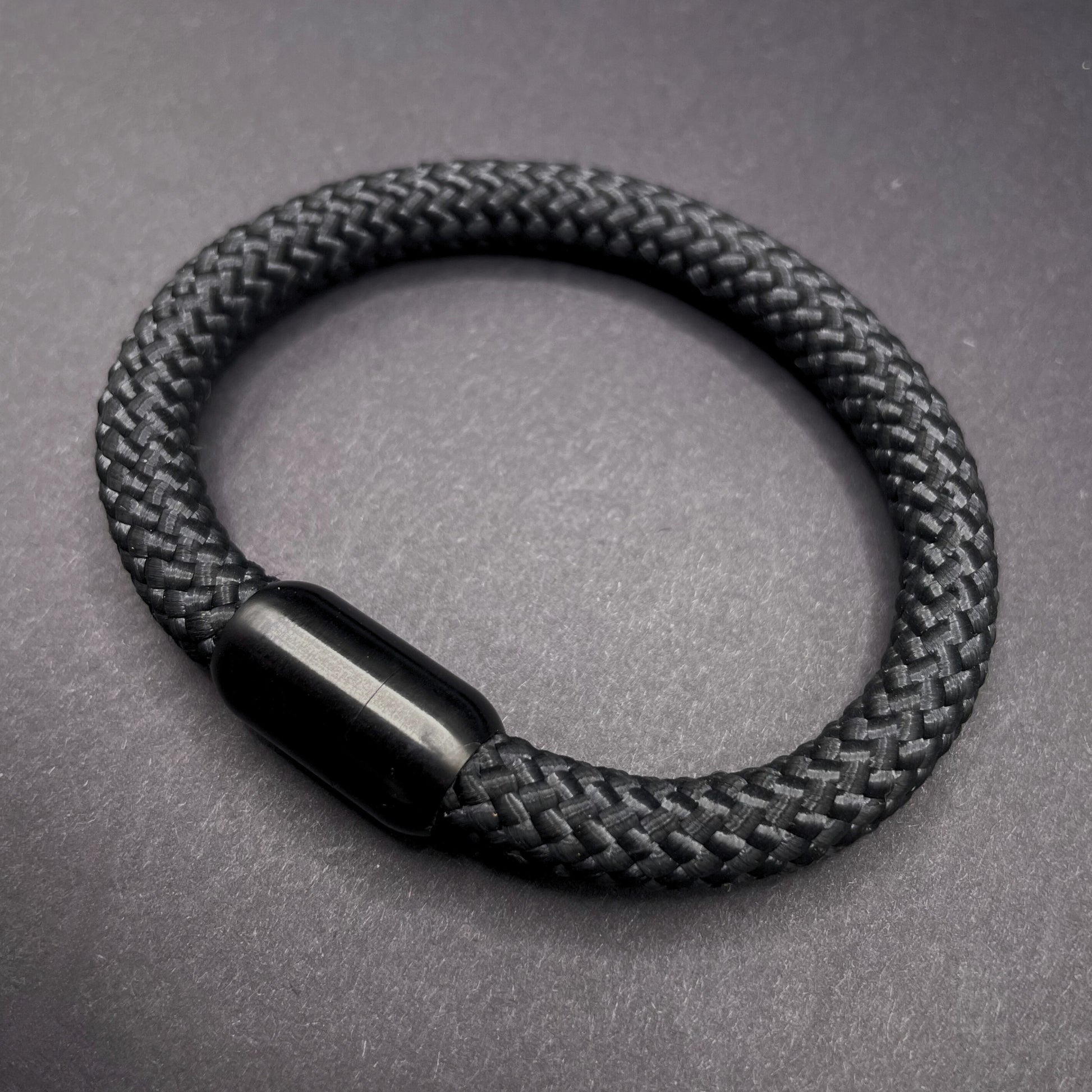 Black braided rope bracelet with a sleek cylindrical black metal clasp, displayed on a dark textured background.