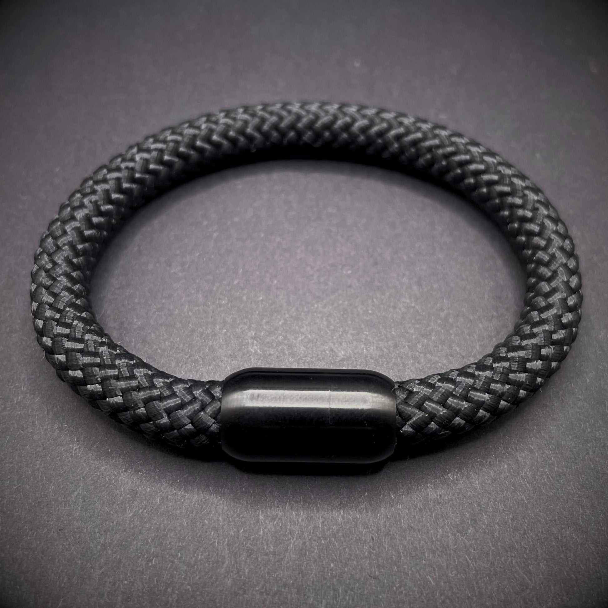 Black braided rope bracelet with a sleek cylindrical black metal clasp, displayed on a dark textured background.