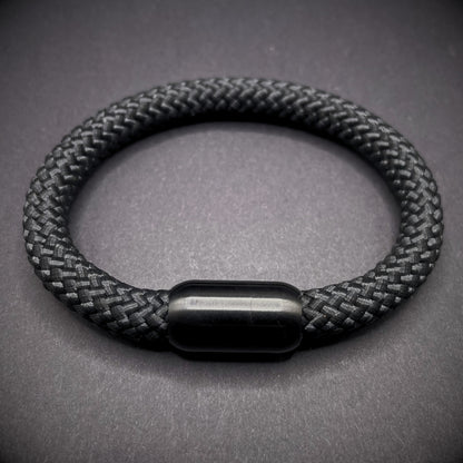 Black braided rope bracelet with a sleek cylindrical black metal clasp, displayed on a dark textured background.