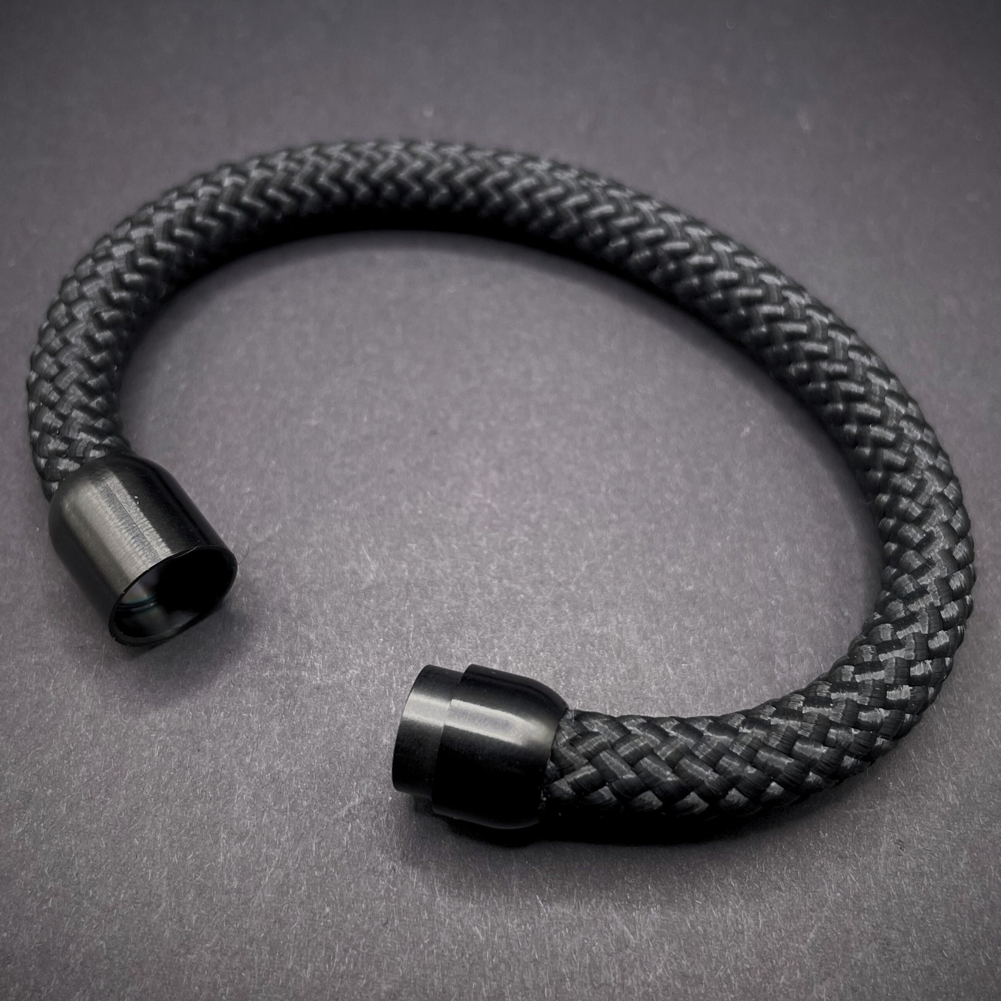 Black braided rope bracelet with a sleek cylindrical black metal clasp, displayed on a dark textured background.