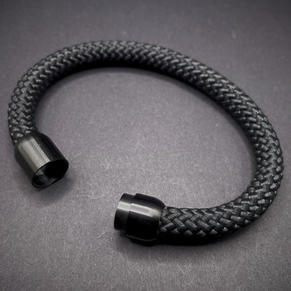 Black braided rope bracelet with a sleek cylindrical black metal clasp, displayed on a dark textured background.