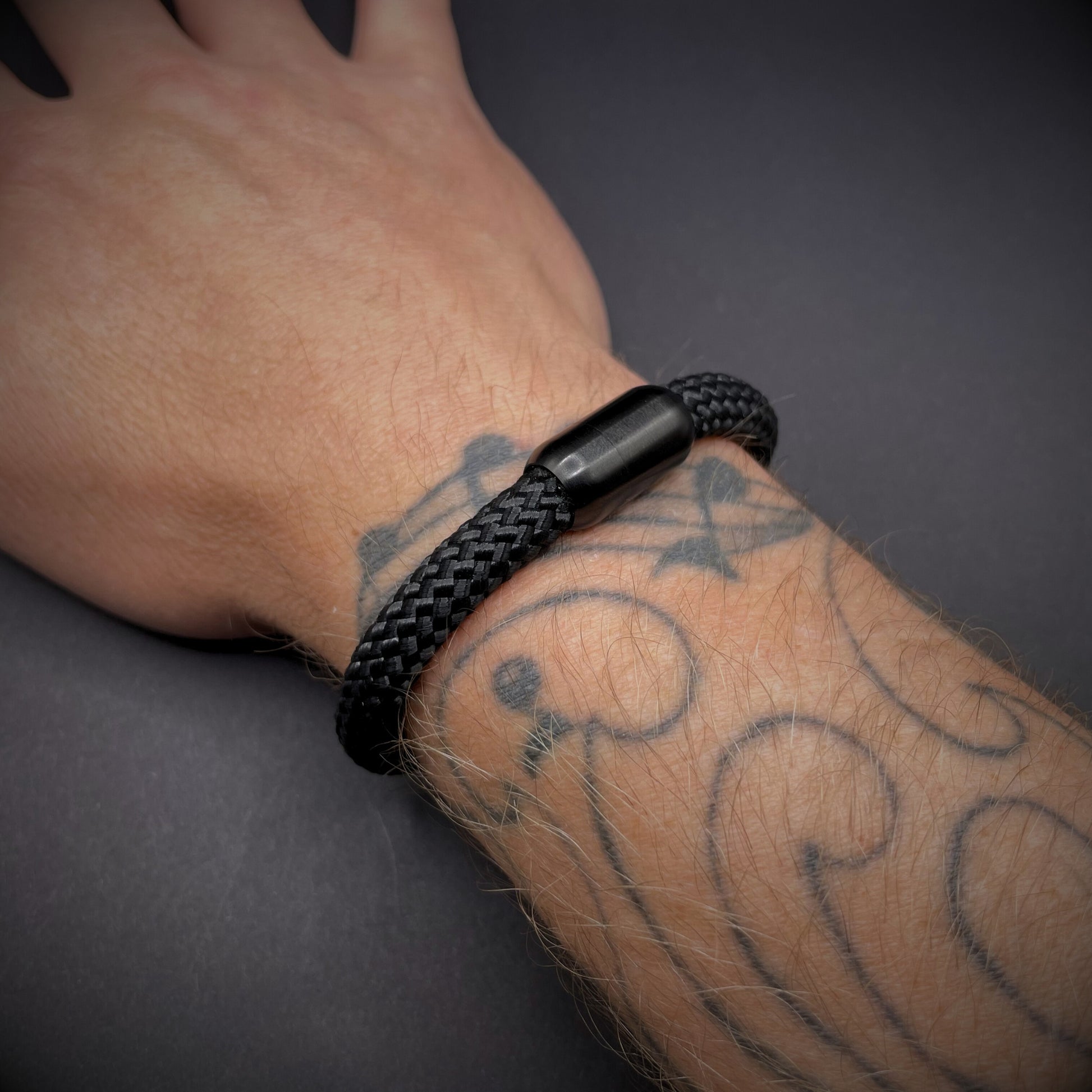Black braided rope bracelet with a sleek cylindrical black metal clasp, displayed on a tattooed wrist over a dark textured background.