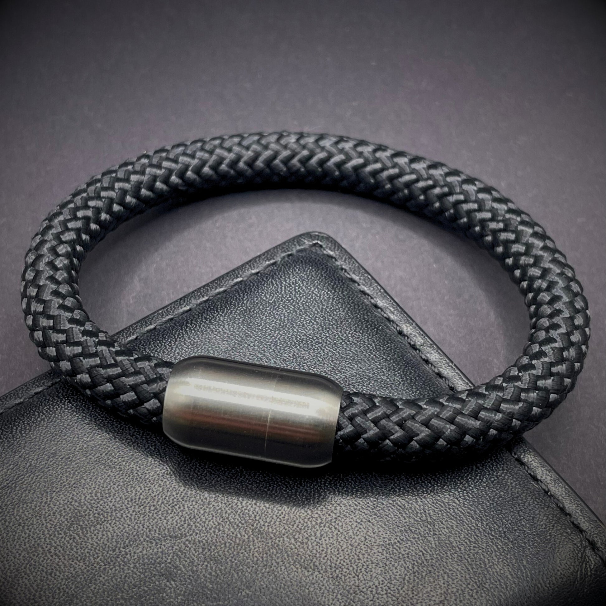 Black braided rope bracelet with a sleek cylindrical brushed steel metal clasp, displayed on a dark textured background.