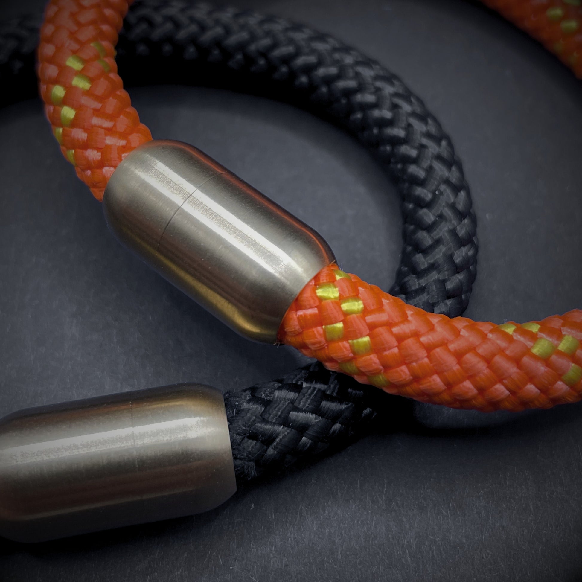 Close-up of two braided rope bracelets, one in bright orange with yellow accents and the other in black, both featuring cylindrical metal clasps, displayed on a dark background.