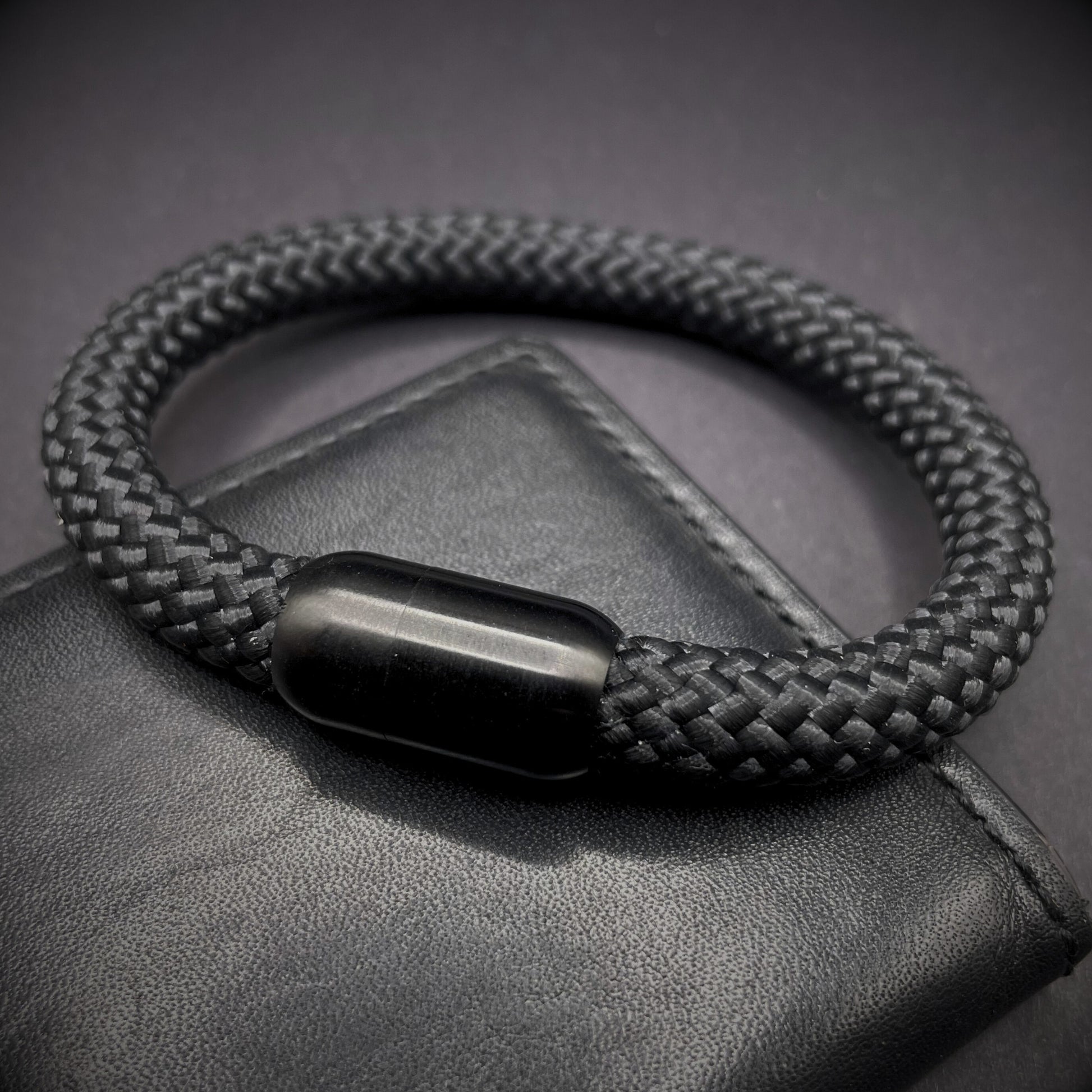 Black braided rope bracelet with a sleek cylindrical black metal clasp, displayed on a dark textured background.