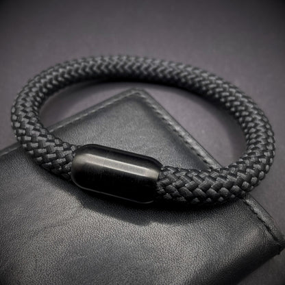 Black braided rope bracelet with a sleek cylindrical black metal clasp, displayed on a dark textured background.