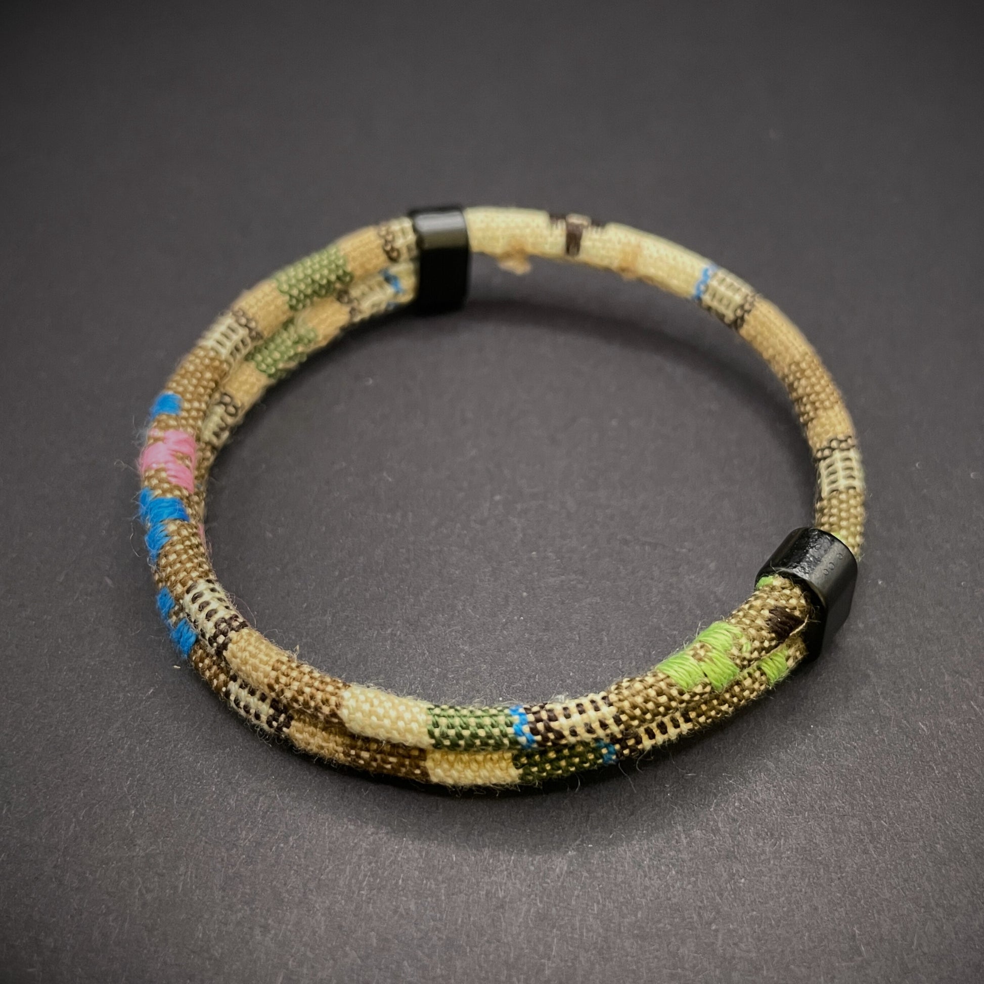 Khaki Boho rope bracelet with black sliding clasps, displayed on a dark grey surface.