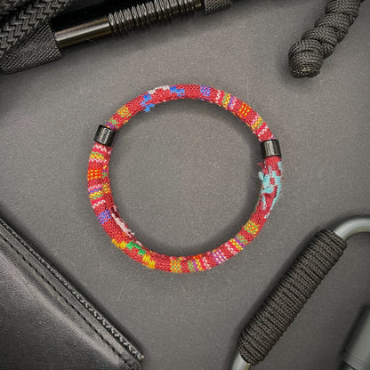 Red Boho rope bracelet with black sliding clasps, displayed on a dark grey surface with EDC gear in the background.